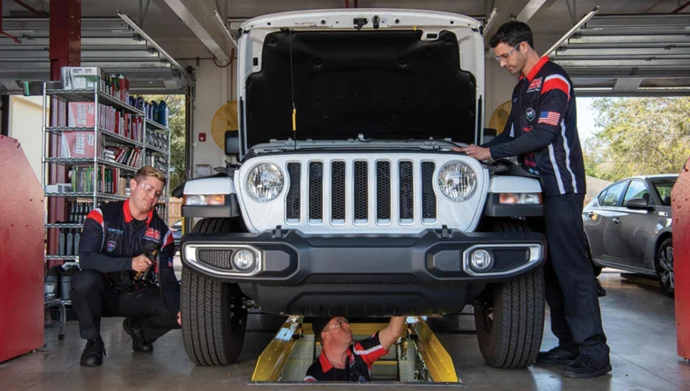 Technicians inspecting a vehicle with the hood raised, including one working underneath and one checking tire pressure